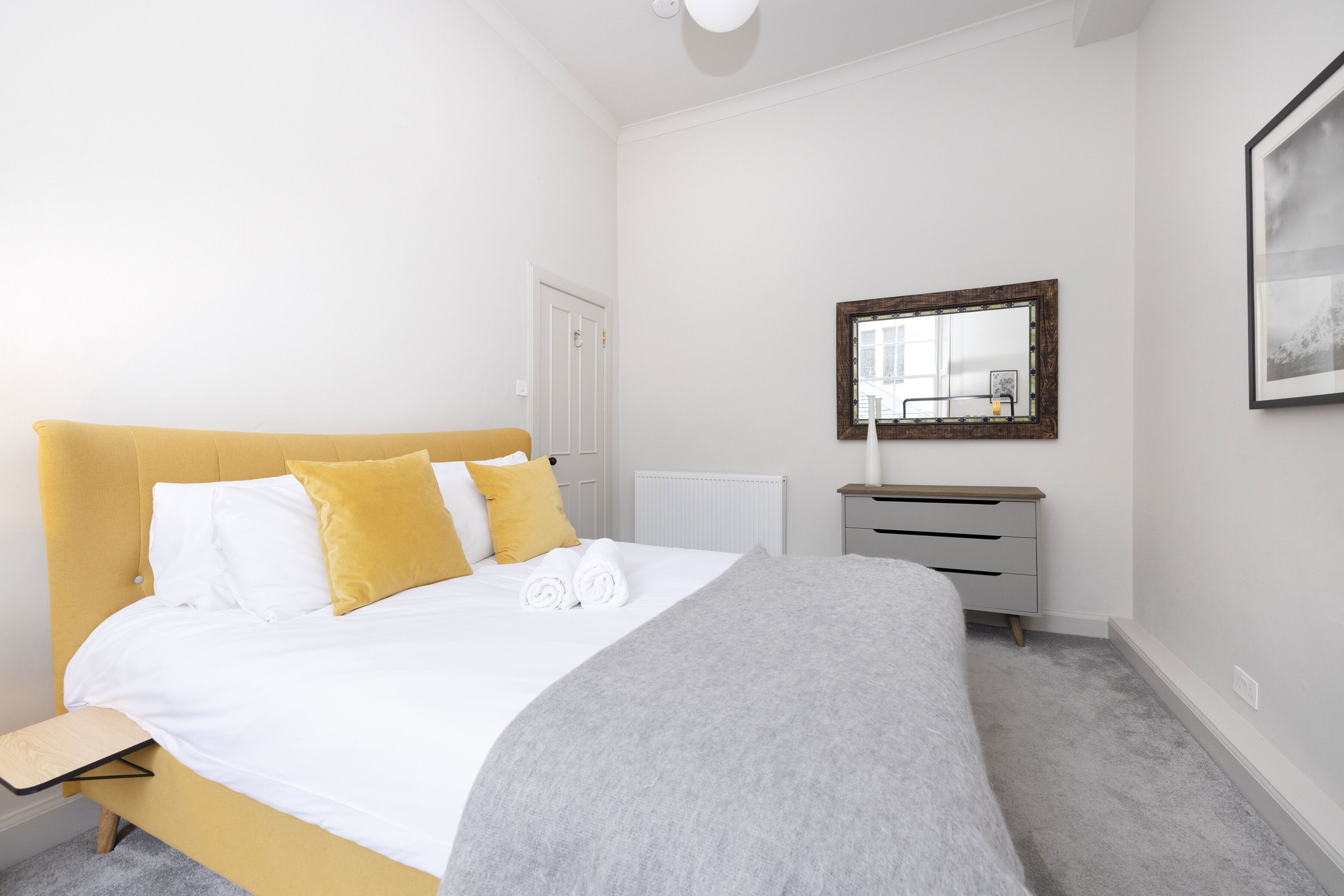 A neatly made bed with white and yellow bedding, a grey throw blanket, and a bedside table with a lamp.
