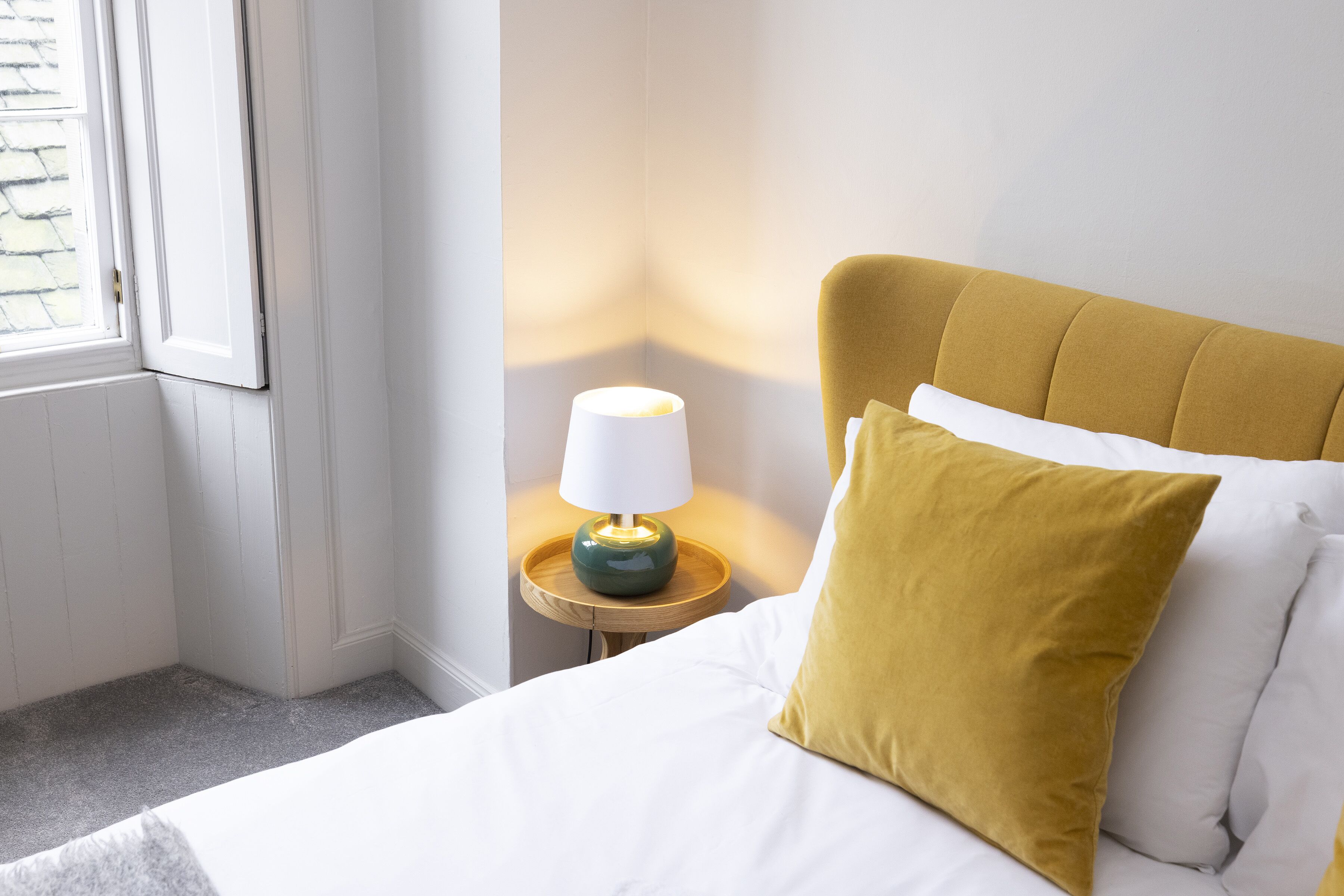 Een netjes opgemaakt bed met witte lakens en een mosterdkleurig kussen, een klein nachtkastje met een lamp, en een raam met zonwering.