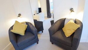 Dos sillones grises con cojines amarillos, una mesita blanca y un mueble blanco en una habitación luminosa con paredes blancas y una ventana.