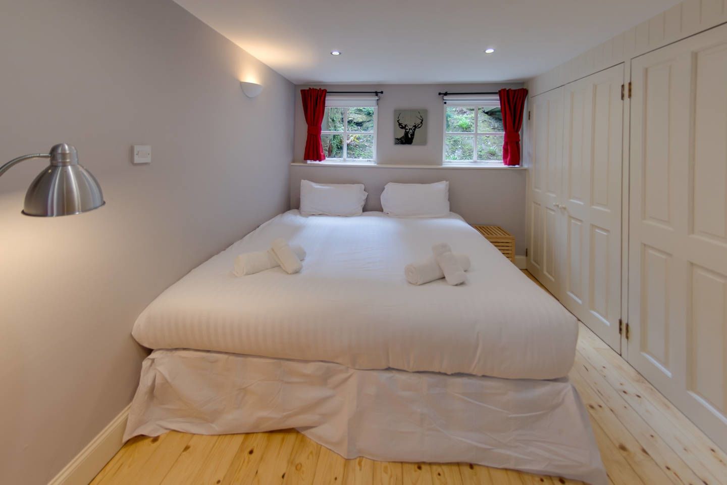 A neatly made bed with white linens and towels, a bedside lamp, and a window with red curtains.