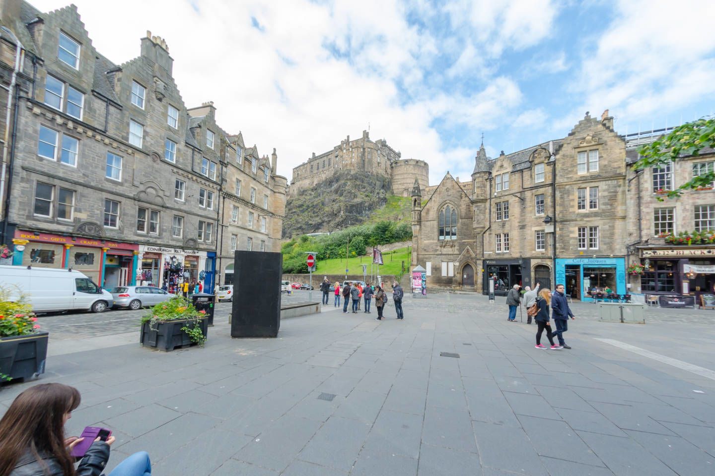 A historic town square with stone buildings, a prominent castle in the background, and a central open space with people and a black sculpture.