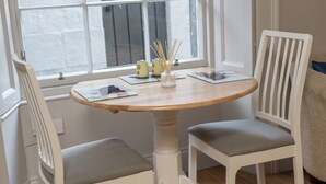 Een ronde houten tafel met twee witte stoelen, een raam met zonwering en een kleine vaas met takken.