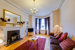 A living room with a fireplace, a television on a stand, a sofa with cushions, a coffee table, and a large mirror above the fireplace. - JOIVY Fabulous Ground-floor 3-bedroom Near Old Town (Edinburgh)