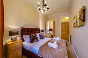 Ein Hotelzimmer mit einem großen Bett, einem Nachttisch mit Lampe, einem Schminktisch, einem Spiegel und einer Chandelier.