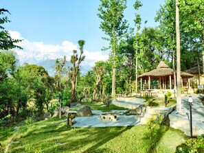A natural setting with a thatched-roof pavilion, stone pathways, and lush greenery. - Dev Bhoomi Farms & Cottages (Dharamshala)