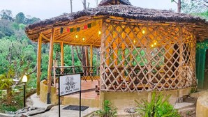 A rustic thatched-roof structure with a lattice roof, surrounded by greenery, and a sign indicating it is part of Dev Bhoom Farms. - Dev Bhoomi Farms & Cottages (Dharamshala)