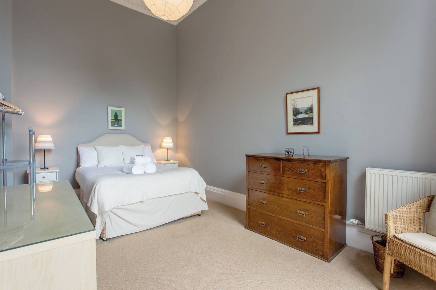 Een slaapkamer met een bed, een houten dressoir, een rieten stoel en een radiator.