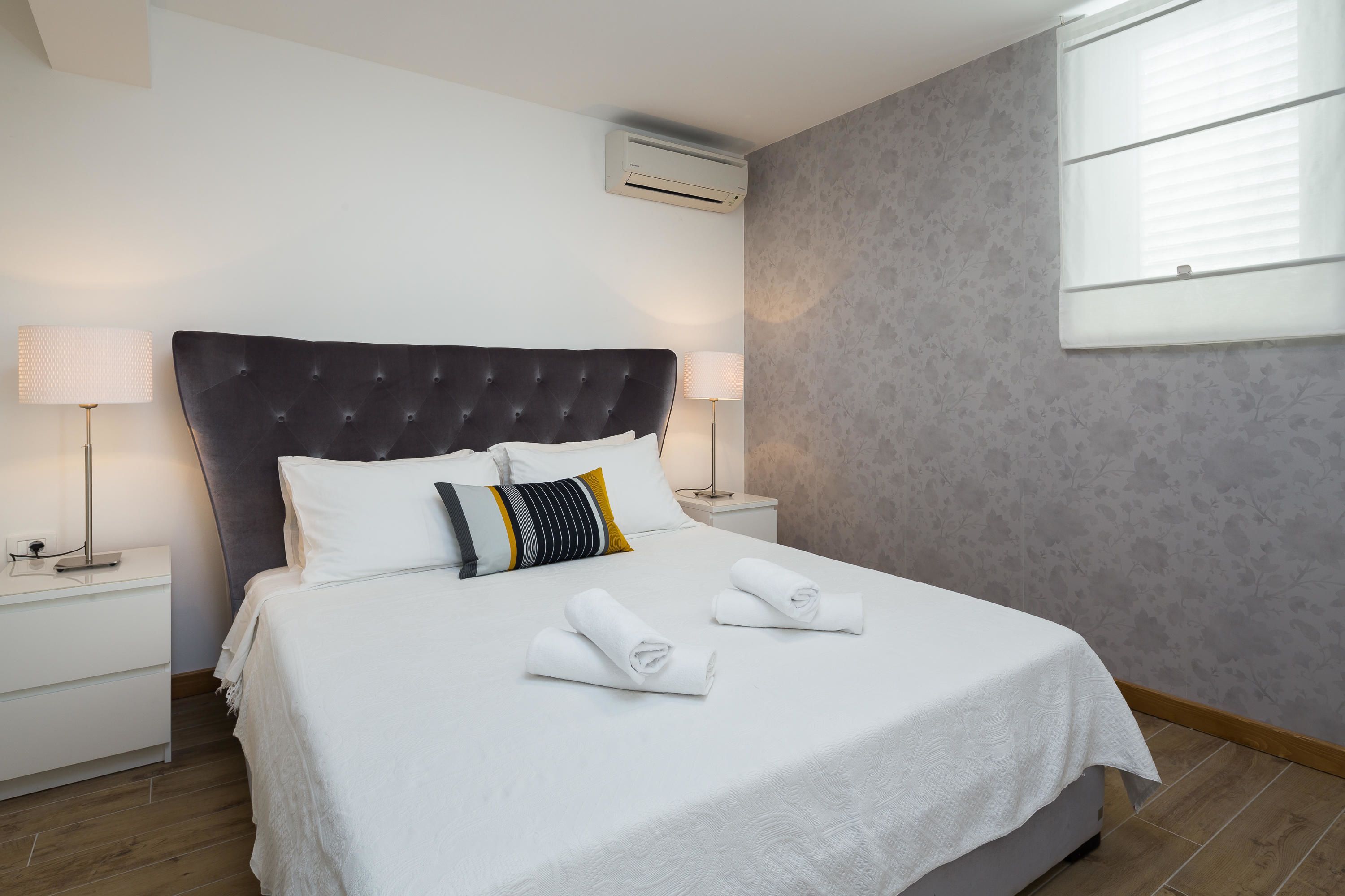 A neatly made bed with white linens and two towels, a dark upholstered headboard, a bedside lamp, and a wall-mounted air conditioner.