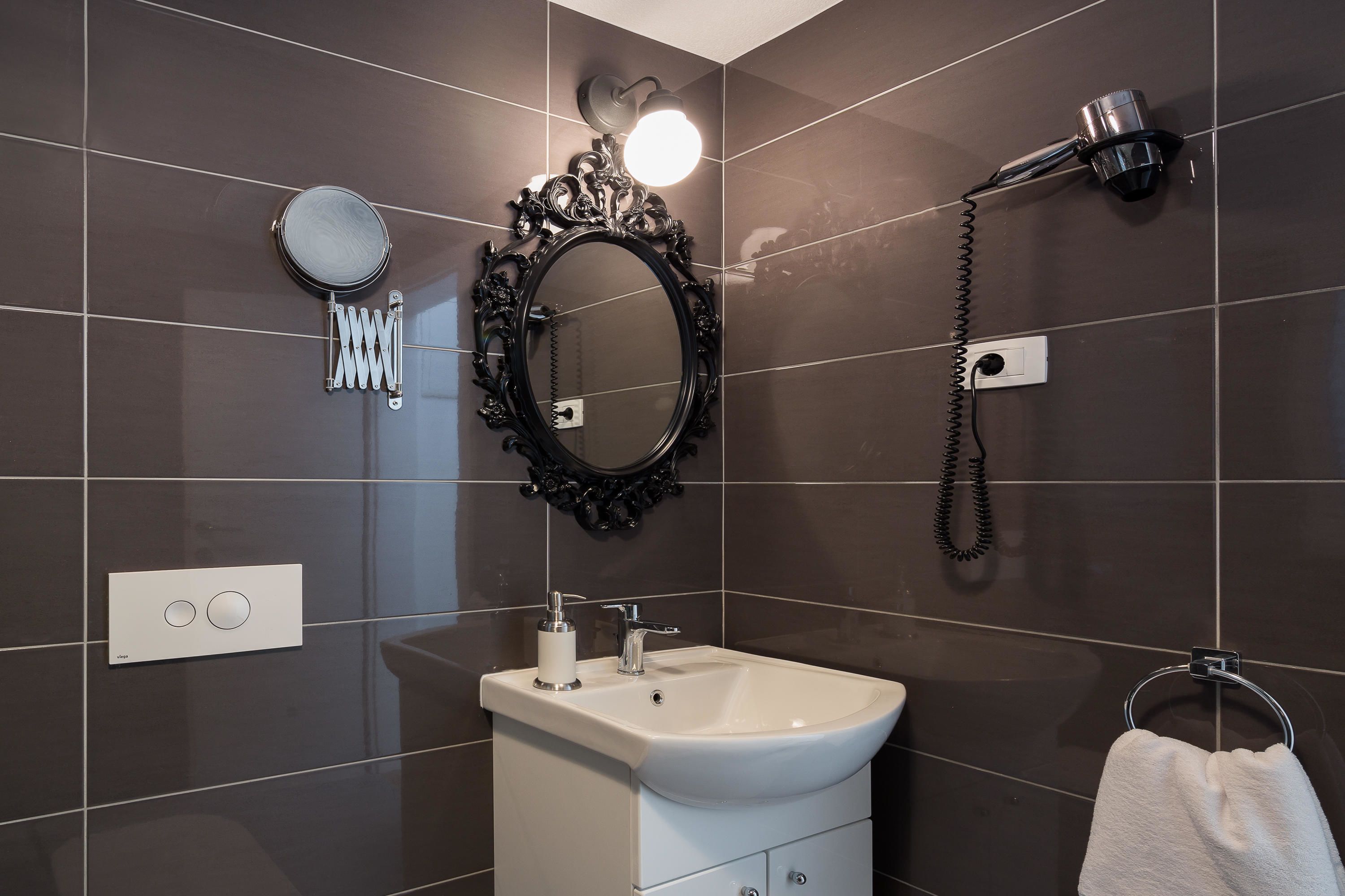 Une salle de bain avec des carreaux de couleur foncée, un lavabo blanc, un miroir doté d’un cadre orné, un sèche-cheveux et un porte-serviettes.