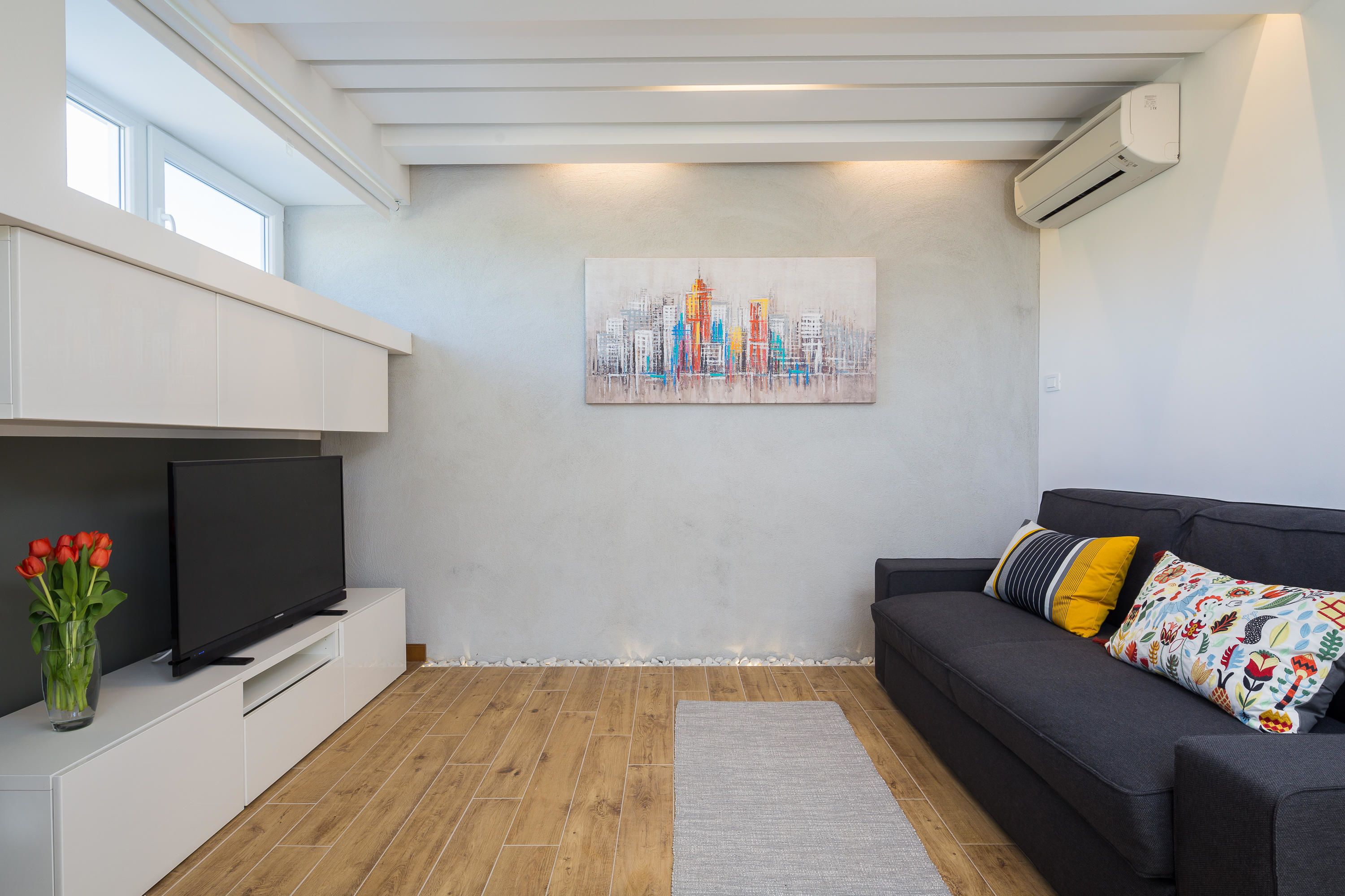 Een moderne woonkamer met een flatscreen televisie, een bank met kleurrijke kussens, een stadsgezicht schilderij aan de muur, en een houten vloer.