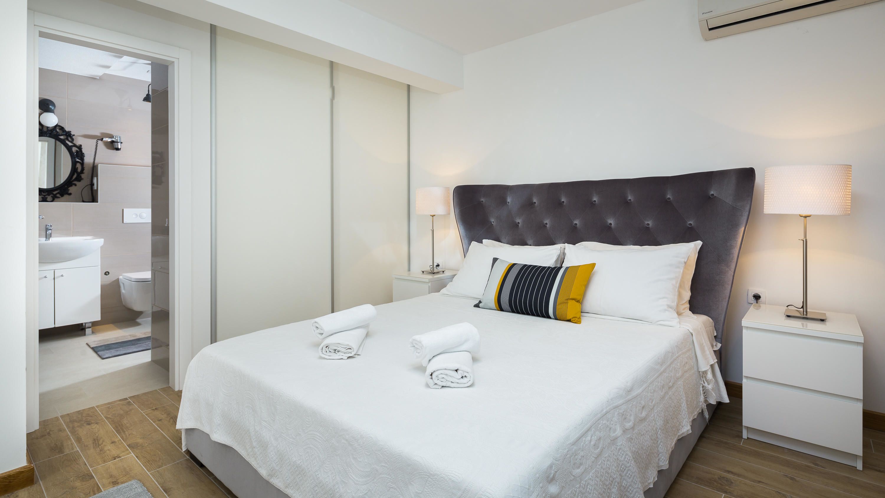 A neatly made bed with white linens and two rolled towels, a dark tufted headboard, and two bedside lamps on white nightstands.