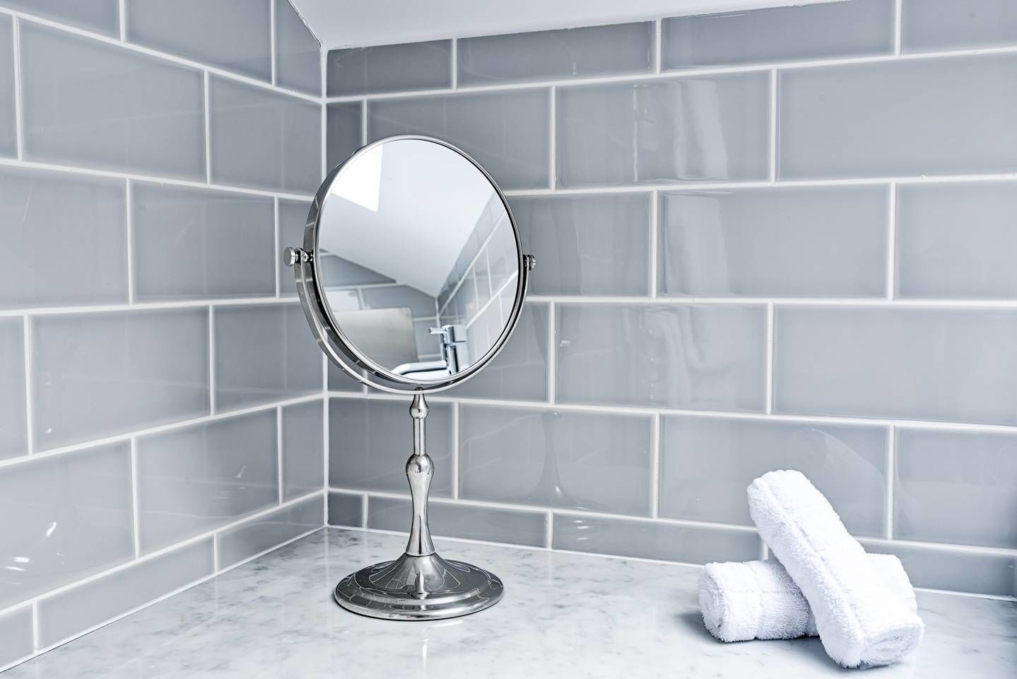 A bathroom with a round mirror on a pedestal, grey tiled walls, and a marble countertop.
