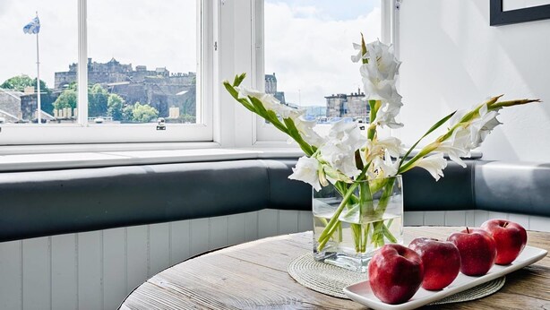 A room with a view of a cityscape through a large window, a vase with white flowers on a table, and a plate of red apples. - JOIVY Luxury George Street Apartments: Castle Suite (Edinburgh)