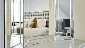 A modern bedroom with a tufted headboard, a bed with white bedding and patterned pillows, a television, and a suitcase on a metal stand. - JOIVY Luxury George Street Apartments: Castle Suite (Edinburgh)