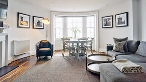 A modern living room with a dining area, featuring a fireplace, a sofa, a coffee table, and framed pictures on the walls. - JOIVY Luxury George Street Apartments: Castle Suite (Edinburgh)