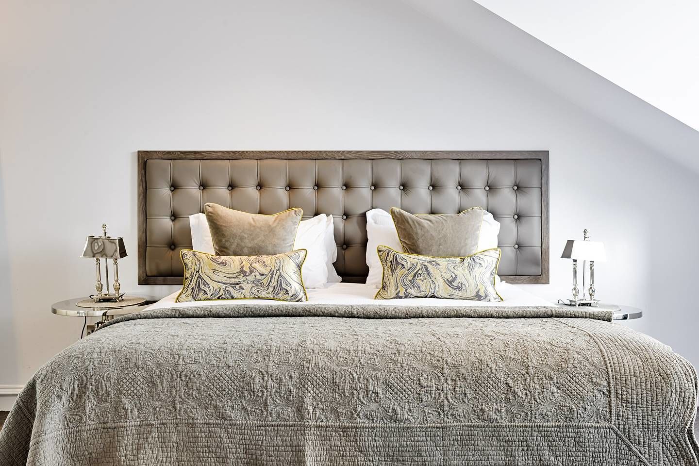 A bedroom with a tufted headboard, a bed with patterned bedding, two bedside tables with lamps, and a view of the sky.