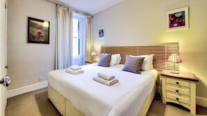 Quarto de hotel bem arrumado com uma cama grande, abajures, uma mesinha de cabeceira e uma janela com cortinas.