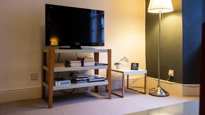 Uma sala de estar com televisão sobre um suporte de madeira, uma mesinha com abajur e uma estante de livros com livros e uma planta.