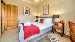 Une chambre à coucher avec un grand lit, une porte en bois, deux œuvres d’art encadrées, un lustre, des lampes de chevet et une commode en bois.