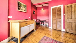 Une pièce avec un plancher en bois, une armoire blanche et en bois, un mur rouge, une porte en bois et une petite table avec des chaises.