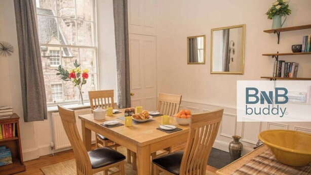 A dining area with a wooden table, four chairs, a bowl of fruit, and a coffee pot. There are bookshelves and a window with curtains. - JOIVY The Old Town Sugar House - Amazing Location (Edinburgh)