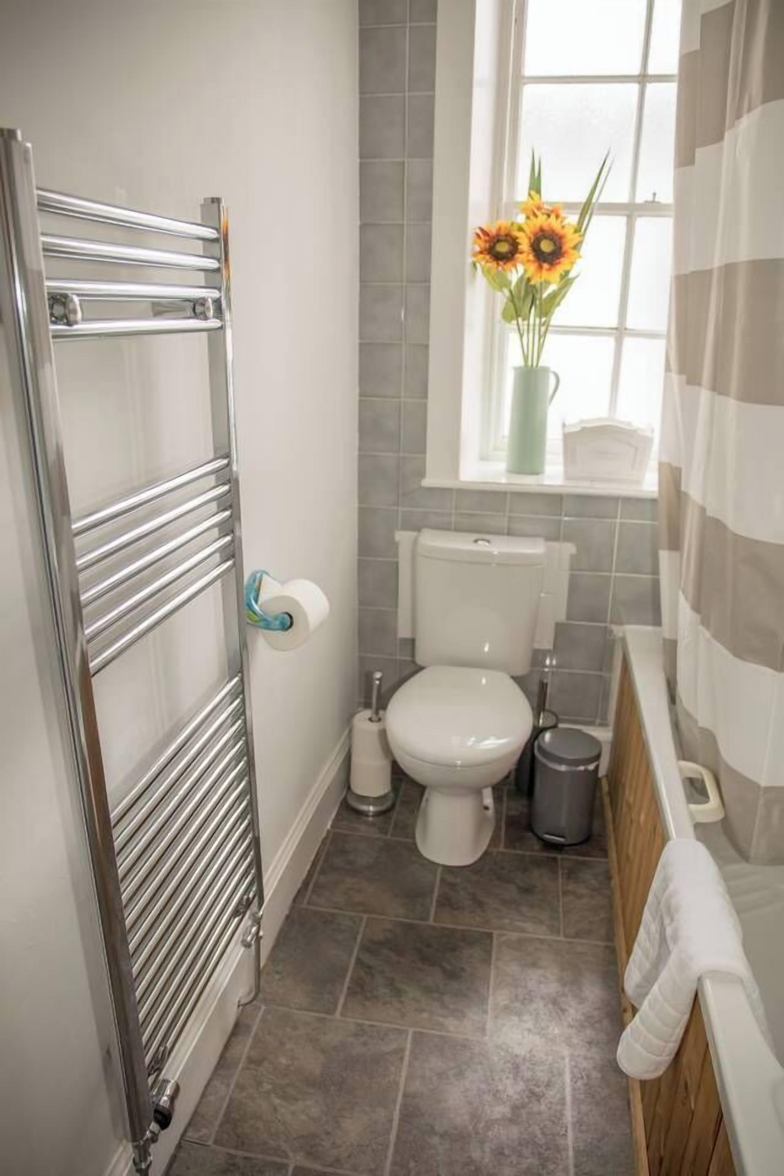 A bathroom with a chrome towel rack, a toilet, a trash bin, and a window with a view of the outside.