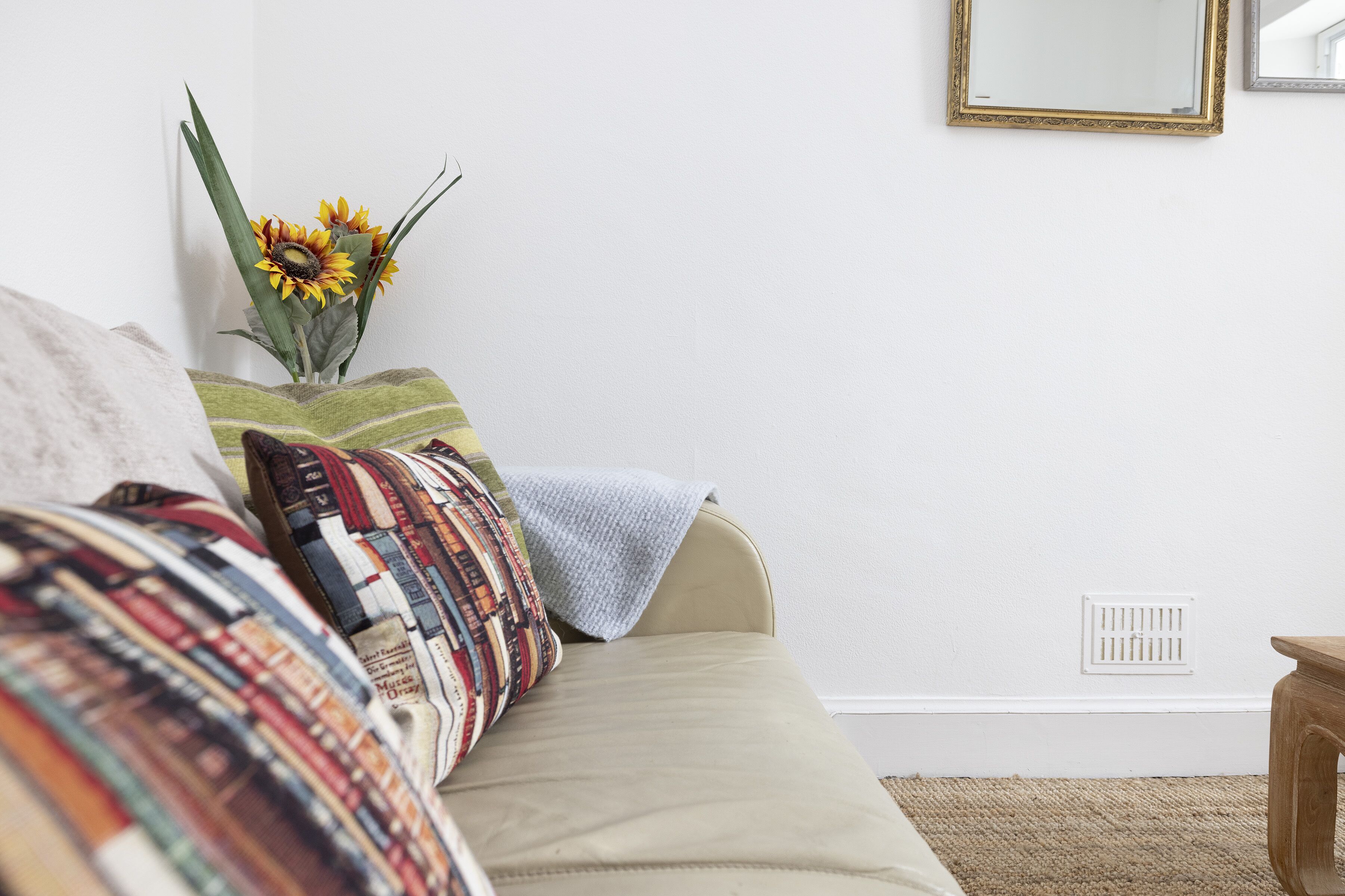 A room with a beige sofa, a patterned throw pillow, a vase with sunflowers, and a mirror on the wall.