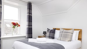 A neatly arranged bedroom with a bed, a bedside table with a lamp, a window with curtains, and a view of the outside. - JOIVY The Old Town Sugar House - Amazing Location (Edinburgh)