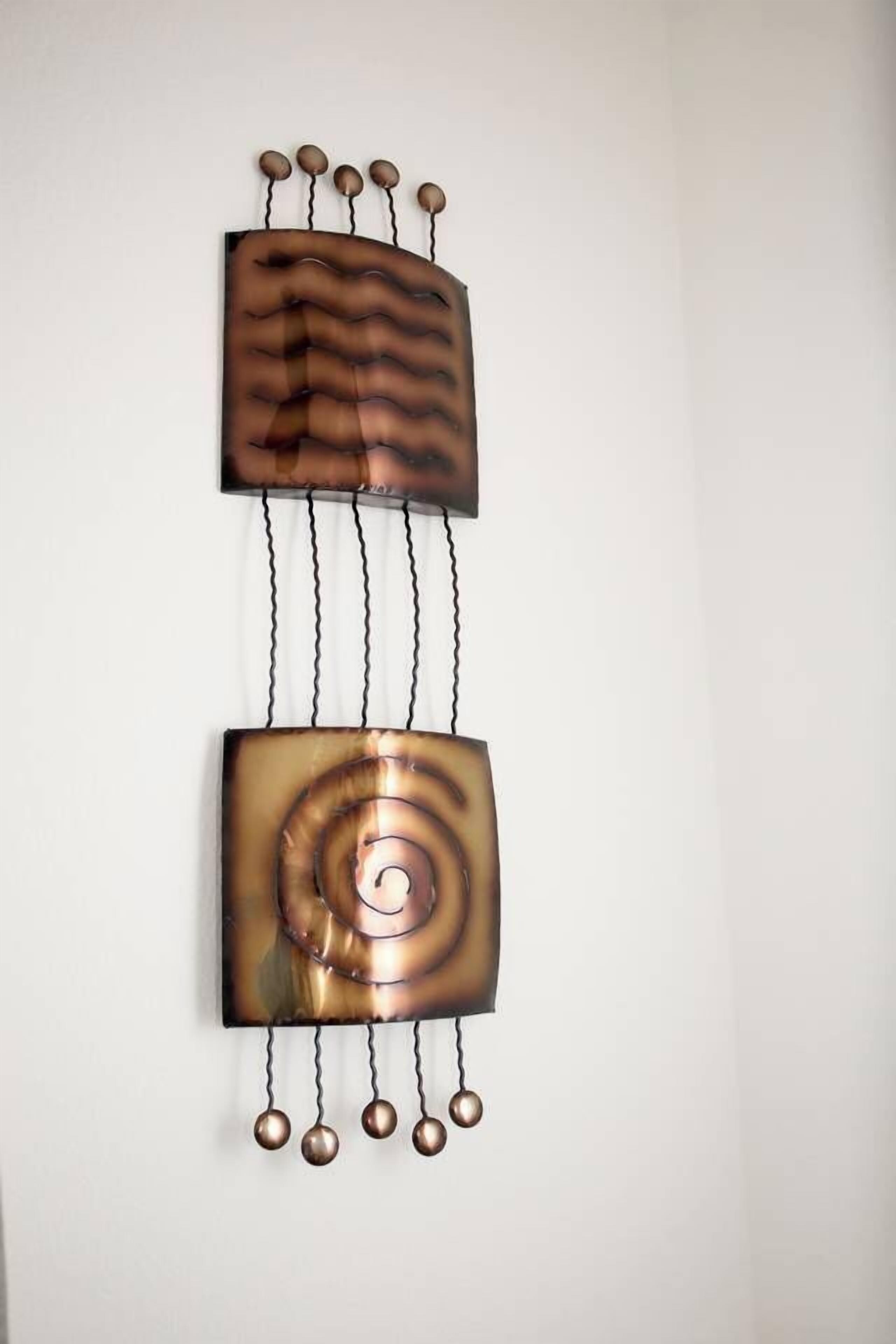 A wall-mounted abstract art piece with a metallic finish, featuring a central spiral design and hanging spherical elements.