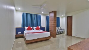 A modern bedroom with a wooden bed, blue curtains, a ceiling fan, and a wooden door. - Hotel Kuber by Sky Stays (Dwarka)