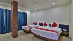 A hotel room with two beds, a wooden column, a ceiling fan, and a large window with blue curtains. - Hotel Kuber by Sky Stays (Dwarka)