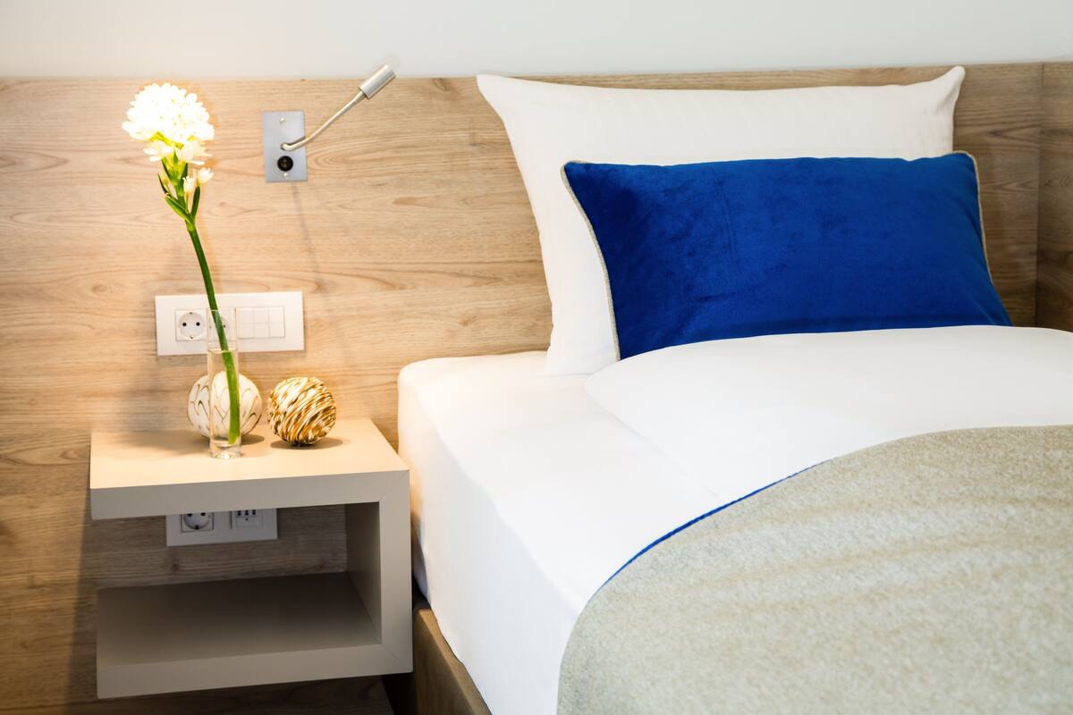 Un lit bien fait, avec une literie blanche et bleue, une table de chevet ornée d’un vase de fleurs et une tête de lit en bois.