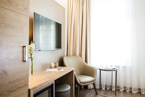 Een moderne hotelkamer met een houten bureau, een comfortabele stoel, een klein rond tafeltje, een flatscreen televisie aan de muur en een raam met dunne gordijnen.