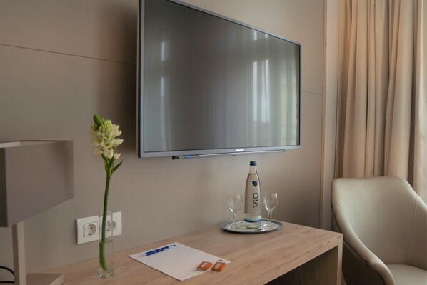 Habitación de hotel con televisor de pantalla plana montado en la pared, escritorio de madera con un florero, una botella de agua, dos vasos, un bolígrafo y una libreta.