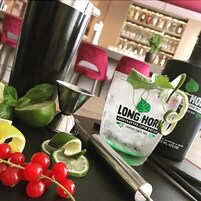 Disposición de bar con un licor artesanal Long Horn, un coctelero y adornos como rodajas de lima y hojas de menta.