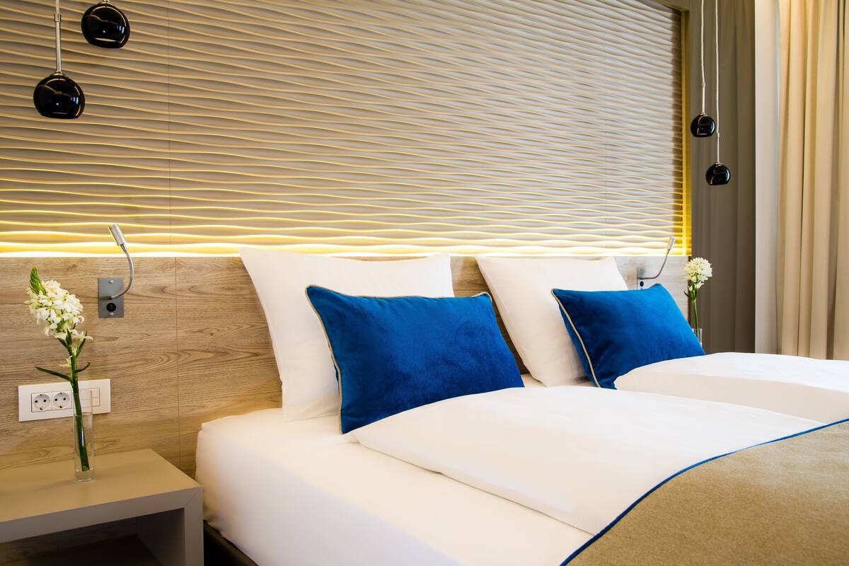Une chambre d’hôtel moderne avec une tête de lit en bois, deux lits avec une literie blanche et des oreillers bleus, une table de chevet ornée d’un vase de fleurs et un luminaire fixé au mur.