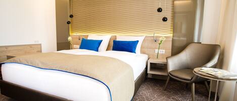 A modern hotel room with a large bed, a curved armchair, a small table, and a wall-mounted lamp.