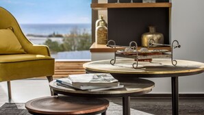 A modern living room with a yellow armchair, a round table with a decorative metal stand, and a view of the ocean through a large window. - Onira Suite Dreams (Hersonissos)