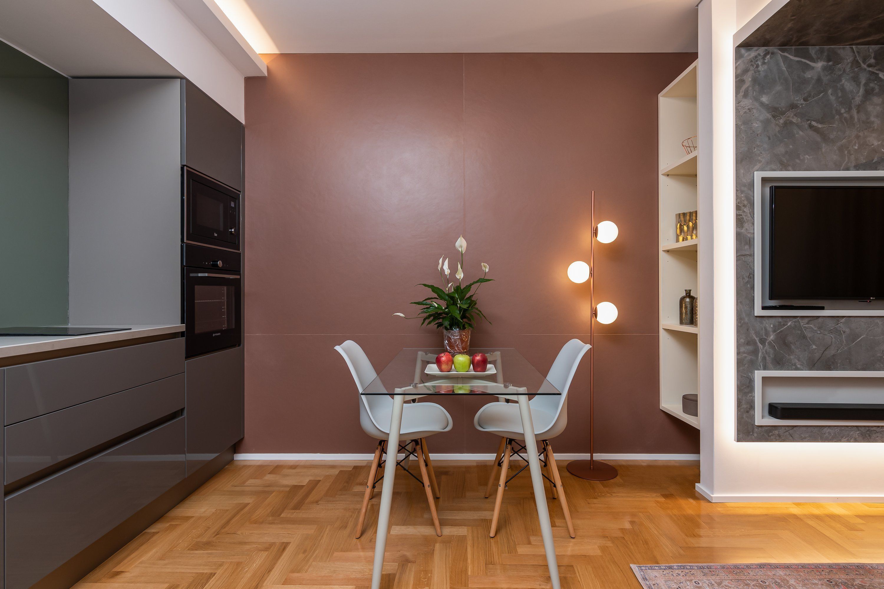 Una cocina moderna con horno y microondas integrados, un comedor con mesa de vidrio y dos sillas, y un televisor montado en la pared.