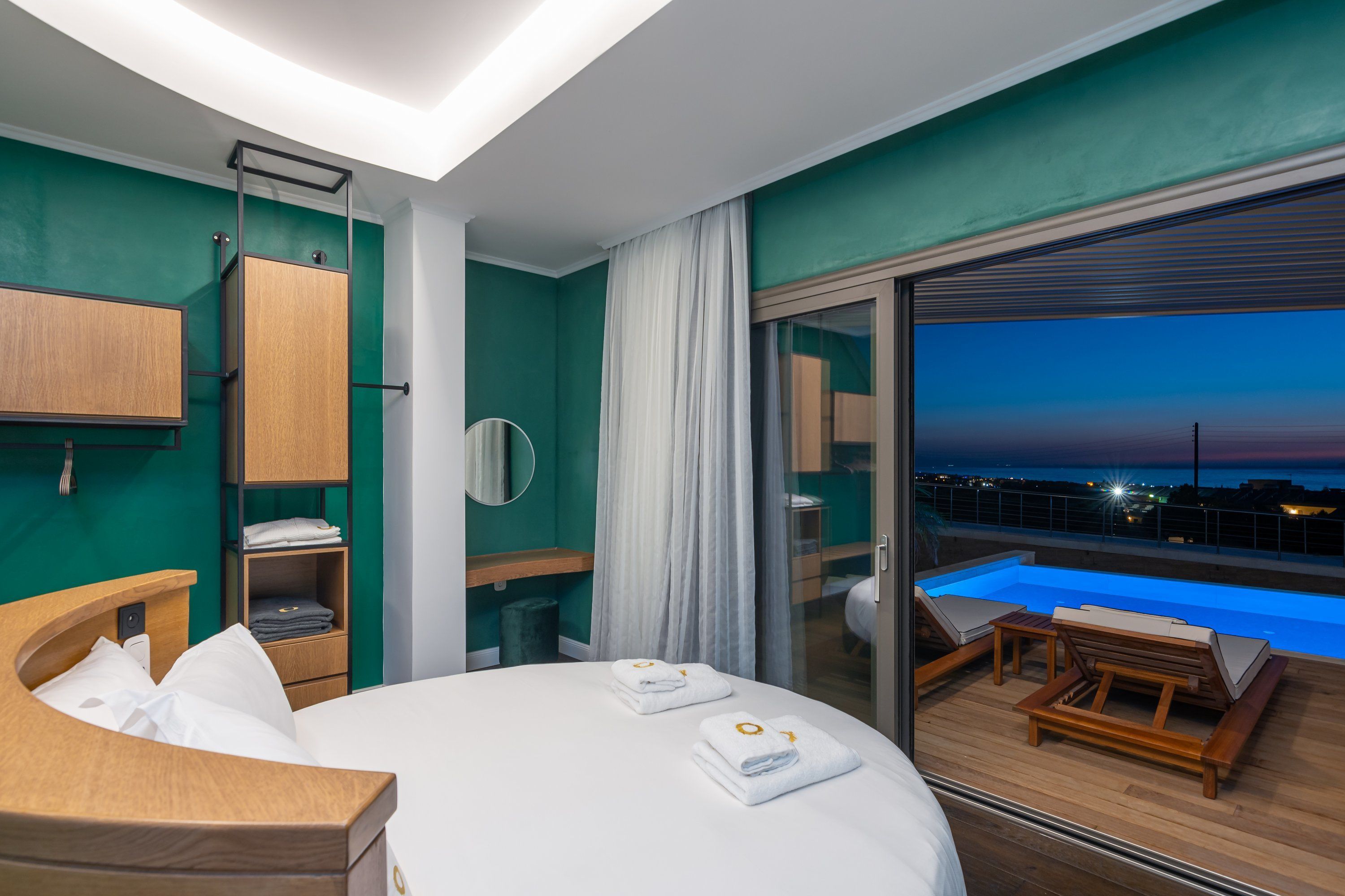 A modern hotel room with a bed, a wooden nightstand, a mirror, a wardrobe, a bathroom with a shower, and a balcony with a pool overlooking a cityscape at dusk.