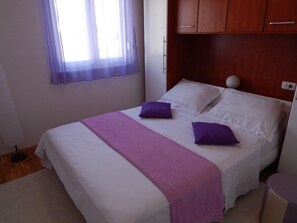 A single bed with white linens and purple pillows, a wooden headboard, a bedside table with a lamp, and a window with a purple curtain.