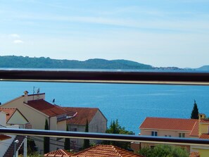 A coastal town with terracotta-roofed buildings, a clear blue sea, and a hilly landscape in the background.