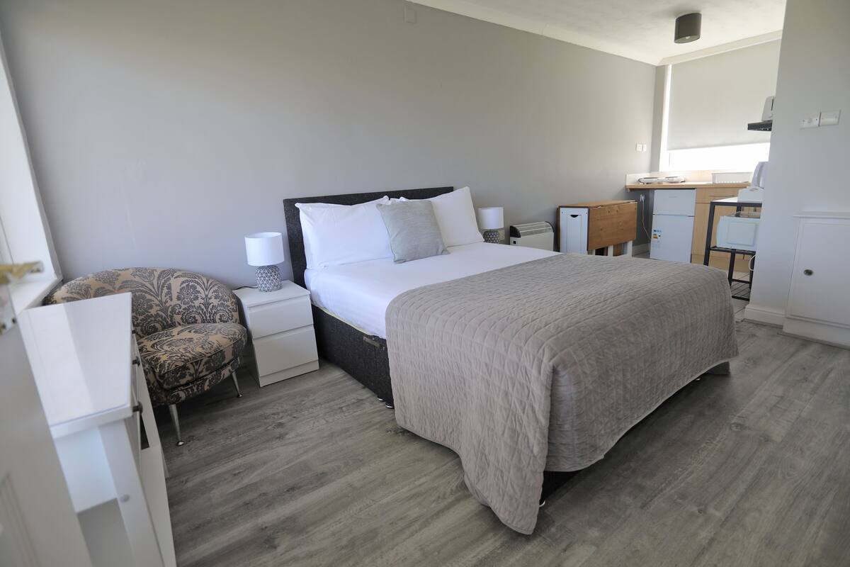 A bedroom with a bed, bedside tables, a chair, a desk, and a kitchenette.