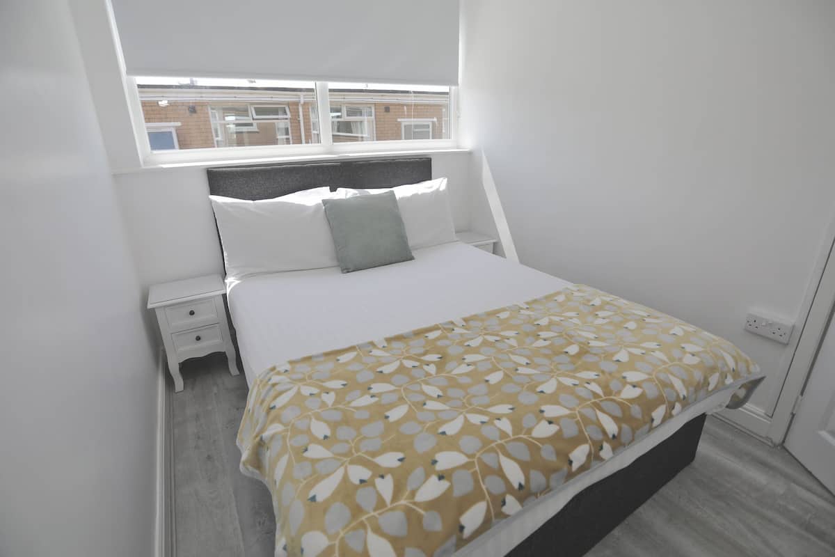 A bedroom with a bed, a nightstand, a window, and a view of buildings outside.