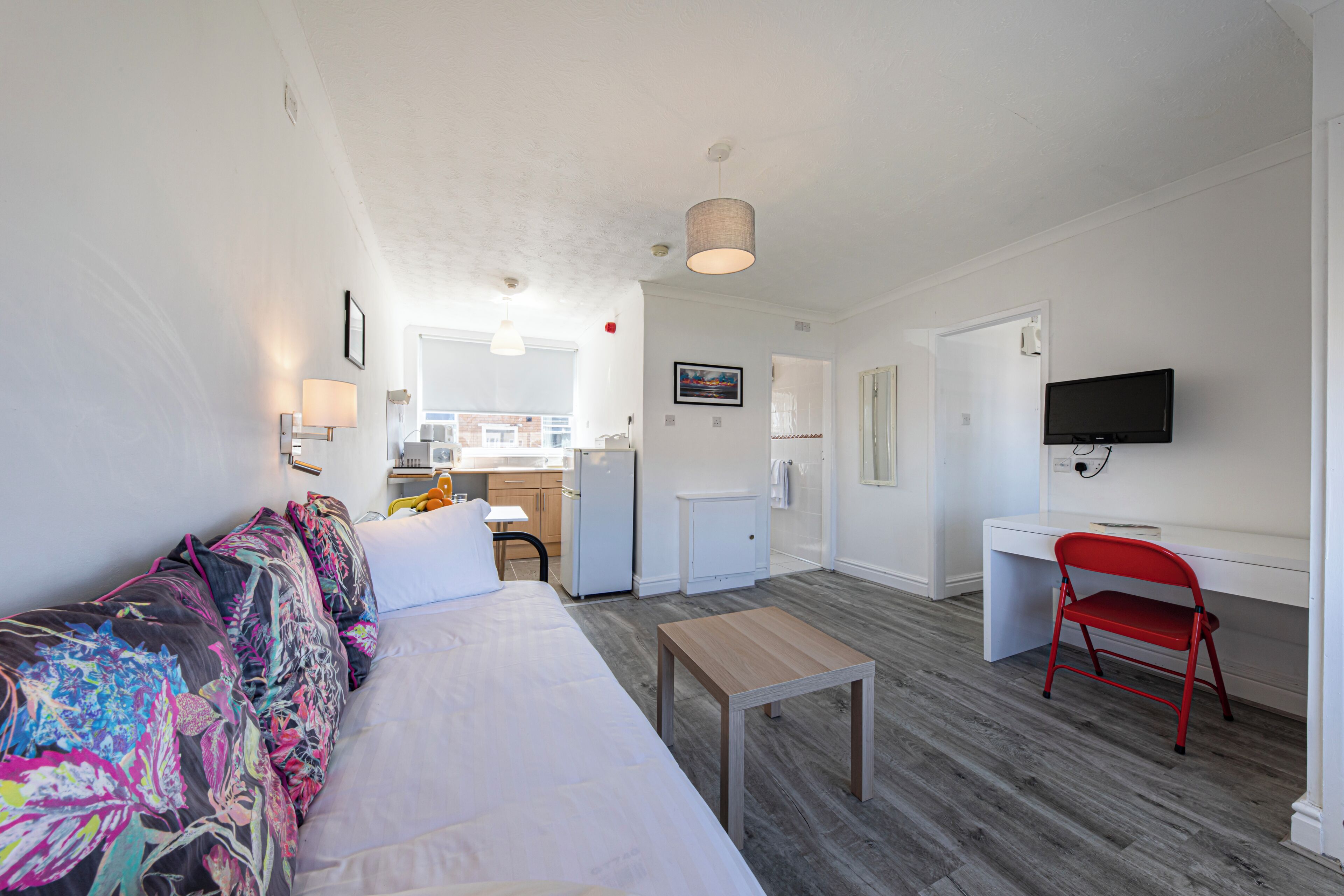 A compact living space with a sofa, a small table, a red chair, a TV, and a kitchenette in the background.