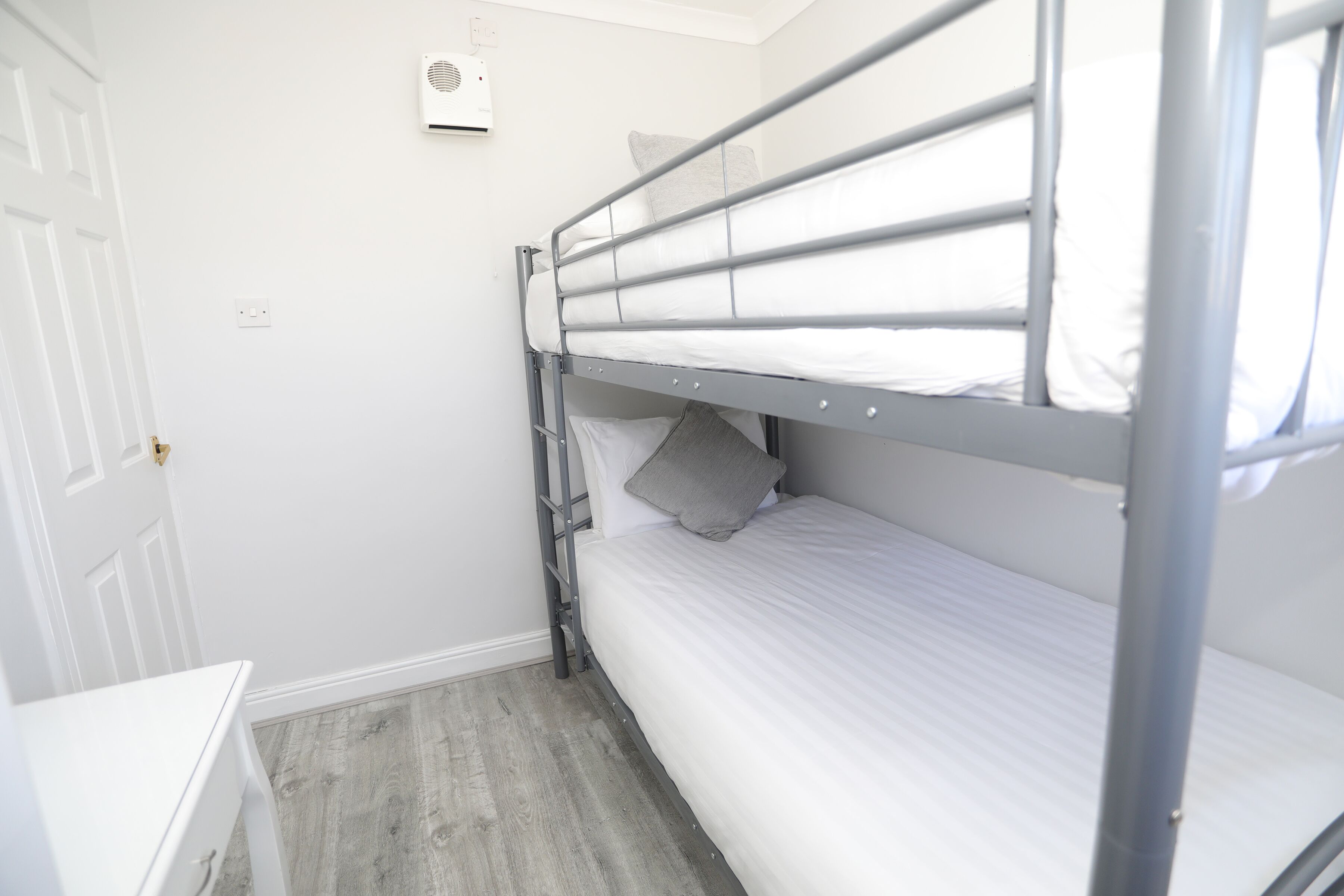 Una cama litera con escalera, un aparador blanco y una puerta en una habitación con paredes blancas y suelo de madera.