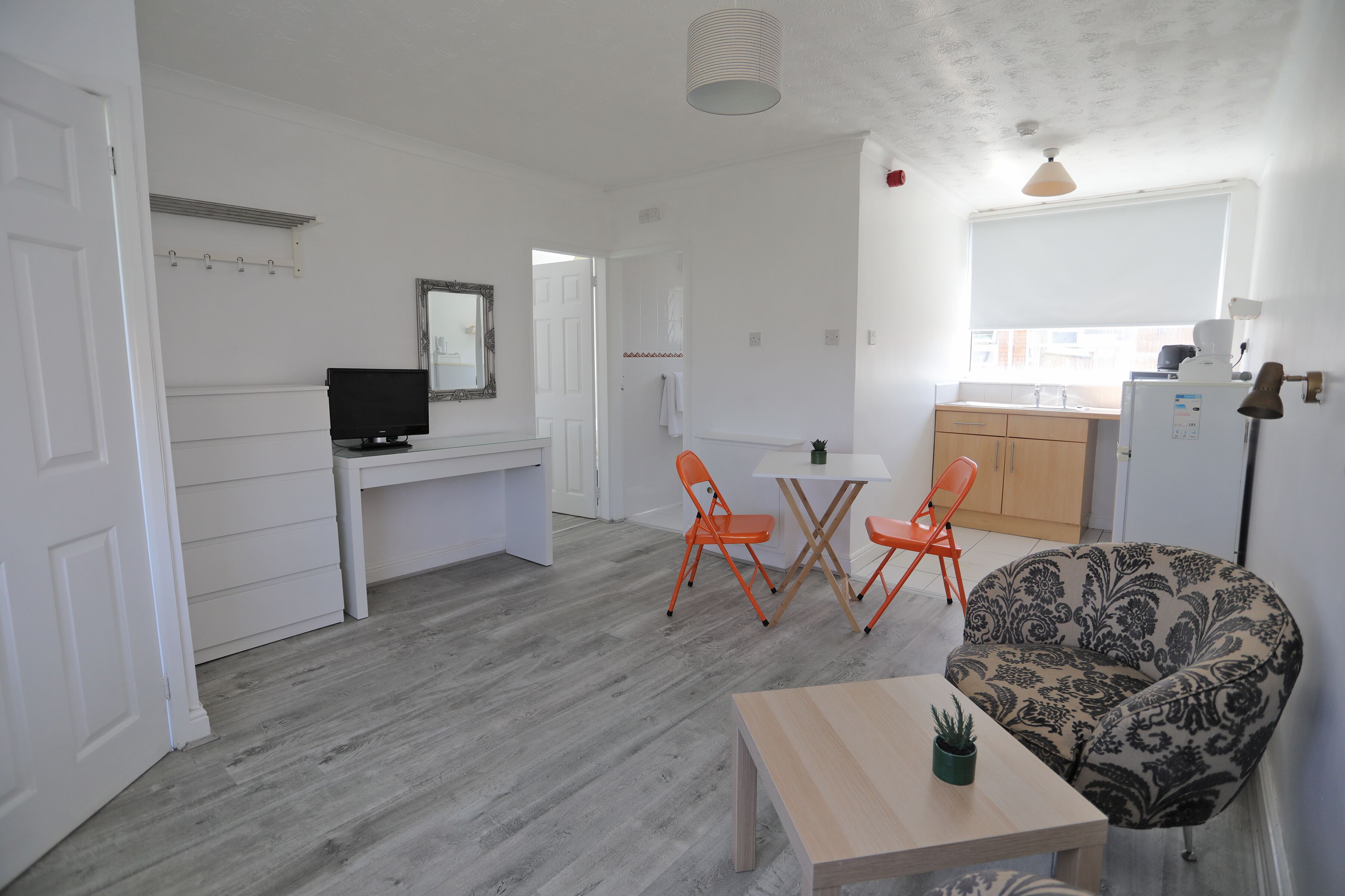 A compact living space with a dining area, a sofa, a small table with a plant, and a kitchen in the background.