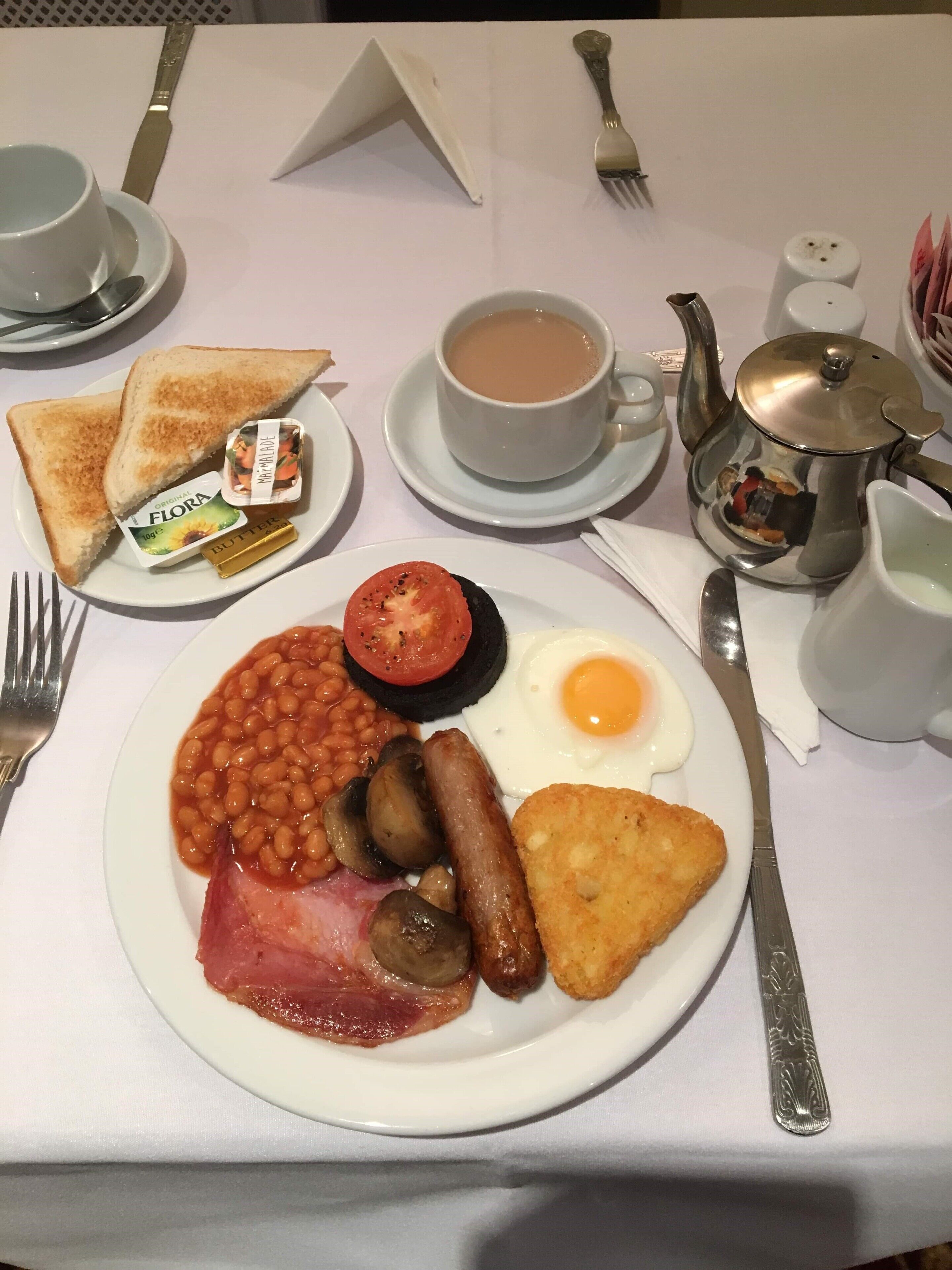 Prato de café da manhã com torradas, manteiga, geleia, tomate, feijão, cogumelos, bacon, linguiça, ovo frito e palha de batata.