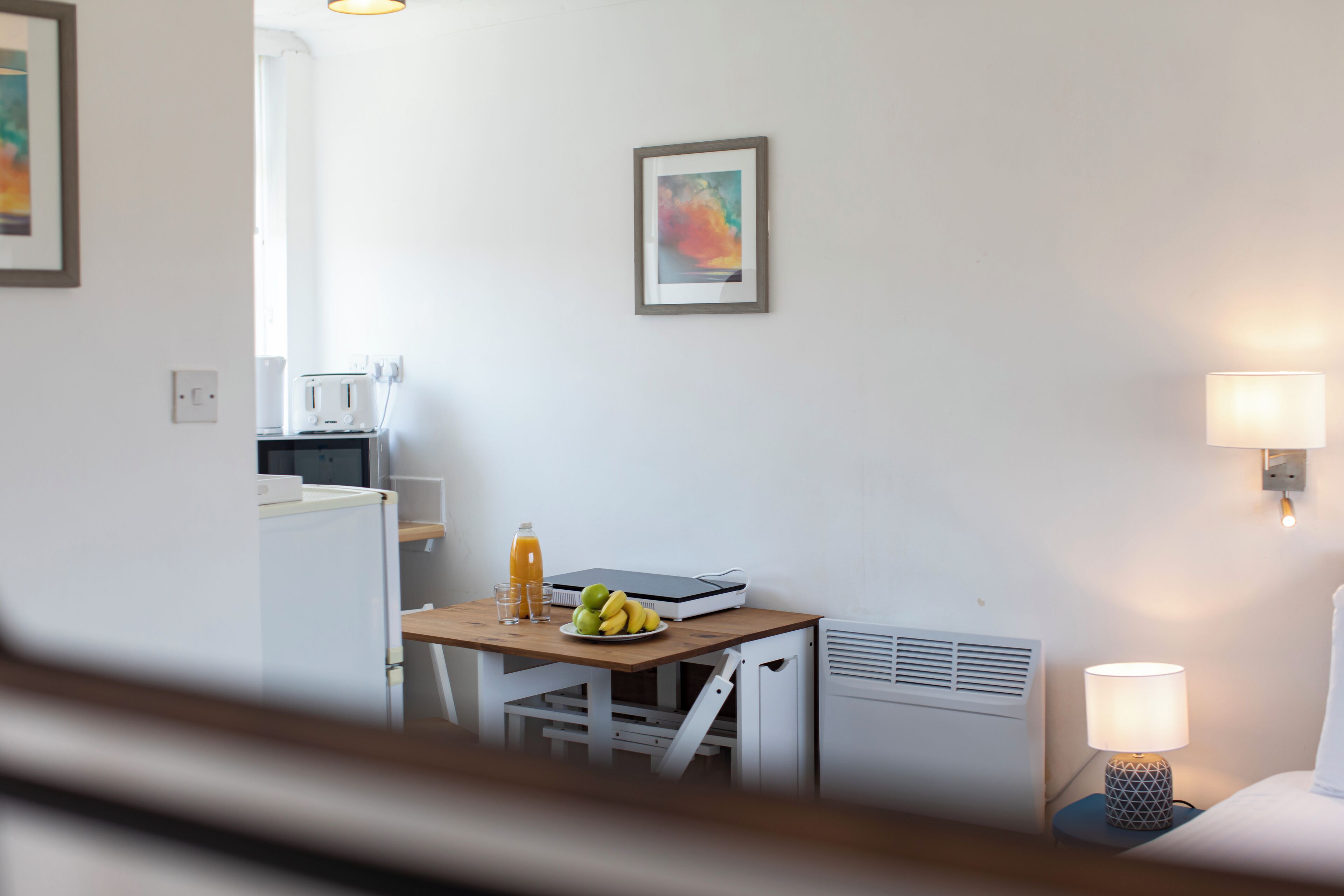 A small kitchenette with a microwave, toaster, and a table with fruits and a juice bottle. There is a framed picture on the wall and a lamp on the bedside table.