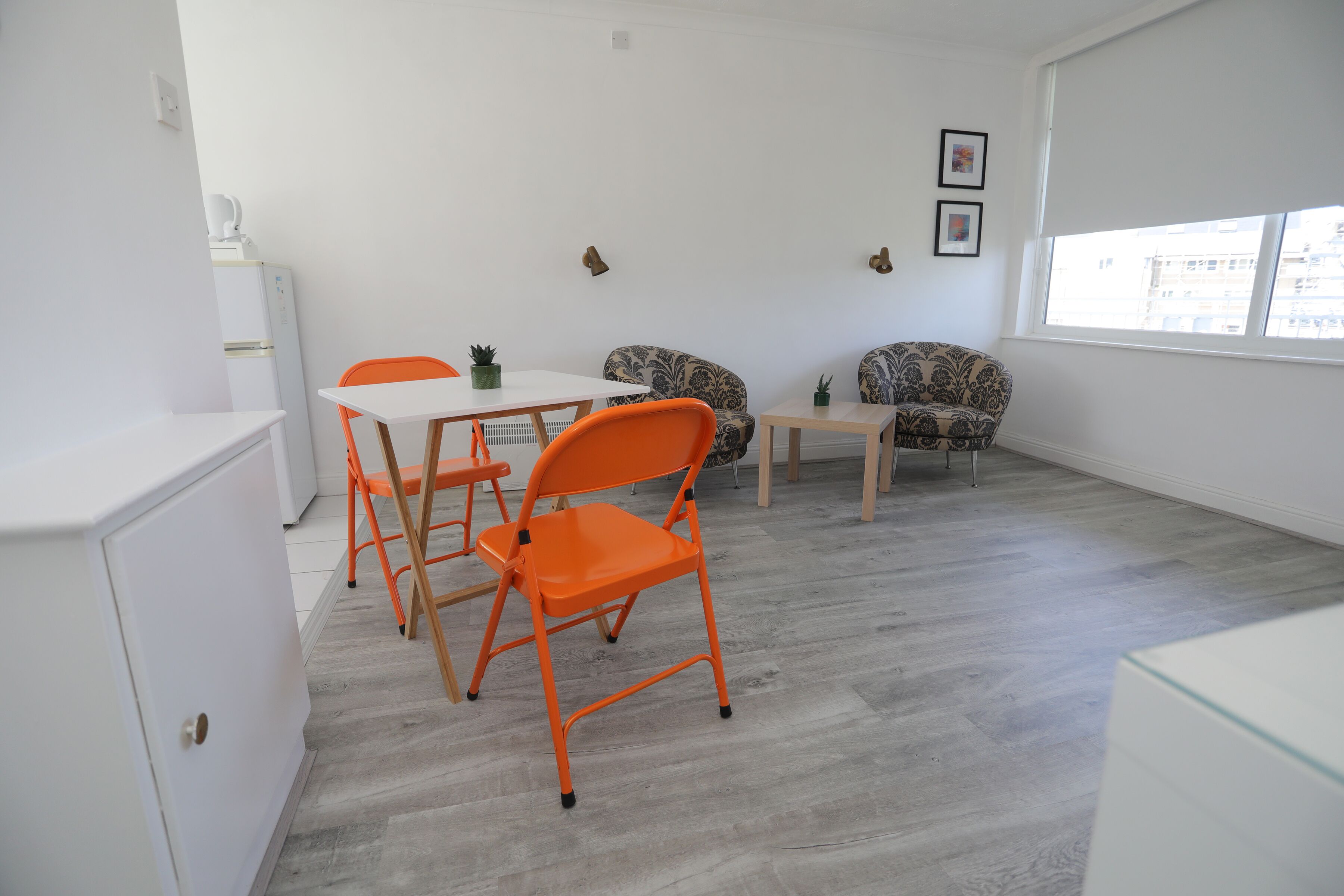 A room with a white cabinet, a table, orange chairs, a patterned sofa, and a small table with a plant.